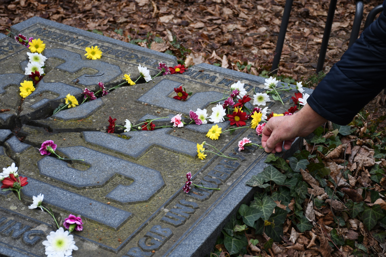 Auf dem Bild sieht man einen Ausschnitt des Gedenksteins und eine Person die eine Blume darauf ablegt. Auf dem Stein sieht man die Jahreszahlen 