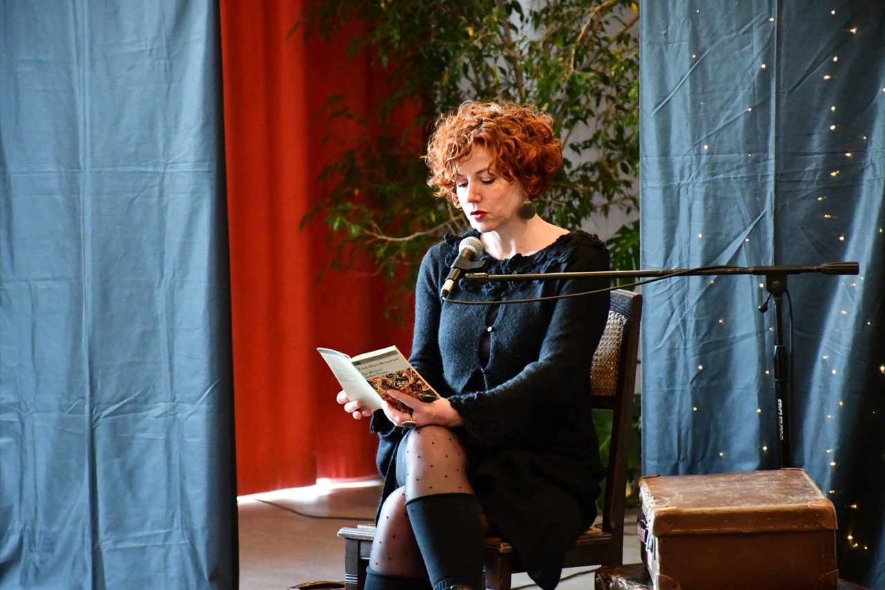 Julia Boegershausen sitzt auf einem Stuhl. In der Hand hält sie ein Buch und spricht in ein Mikrofon.